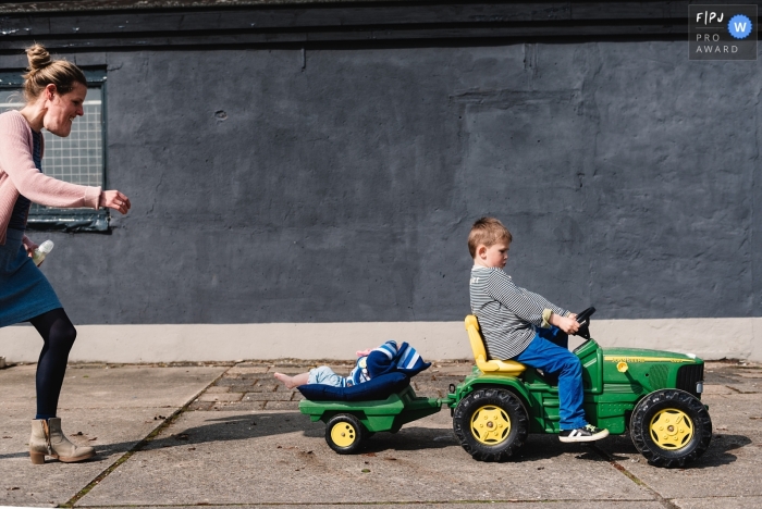 Un garçon conduit le tracteur d'un enfant avec son petit frère à l'arrière alors que sa mère le suit sur cette photographie d'un photographe documentaire familial de Gueldre, aux Pays-Bas.