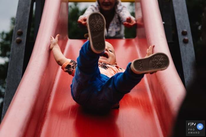 A little boy laughs as he goes down a playground slide in this Family Photojournalist Association awarded photo by a Gelderland, Netherlands documentary family photographer.