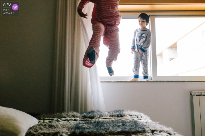 A boy jumps on his bed while his brother stands on a window sill in this photo by an Aveiro, Portugal award-winning family photographer. 