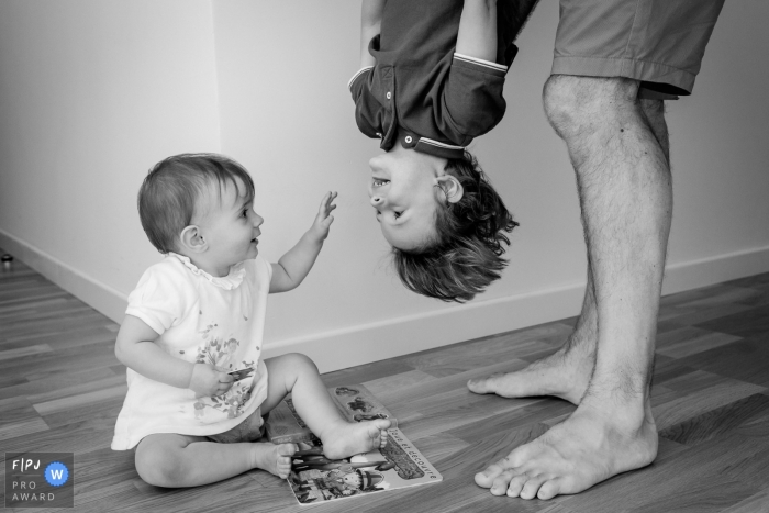 Photo by: <a href="https://www.fpja.com/documentary-family-photography-contests/sybil-rondeau/nantes/2090861">Sybil Rondeau - France</a> A little girl looks at her brother as her father holds him upside down in this Family Photojournalist Association contest awarded photo created by a Nantes family photographer.