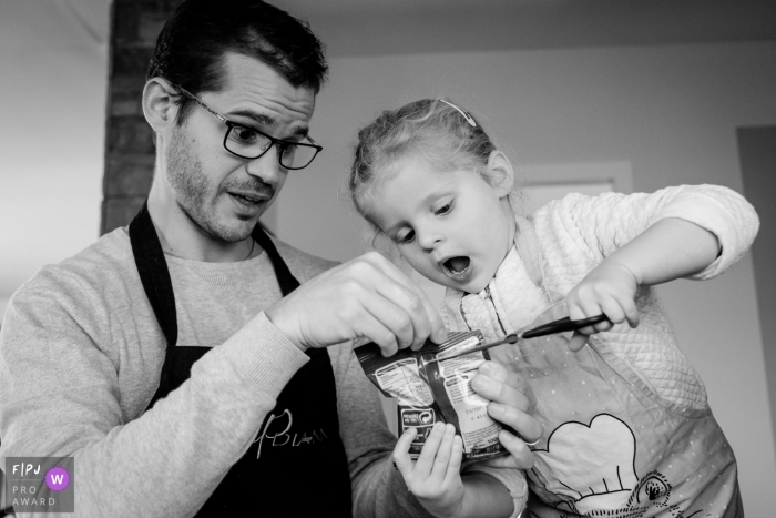 Photo by: <a href="https://www.fpja.com/documentary-family-photography-contests/sybil-rondeau/nantes/2090896">Sybil Rondeau - France</a> A father holds a bag while his daughter cuts it open in this documentary-style family photo captured by a Nantes photographer.