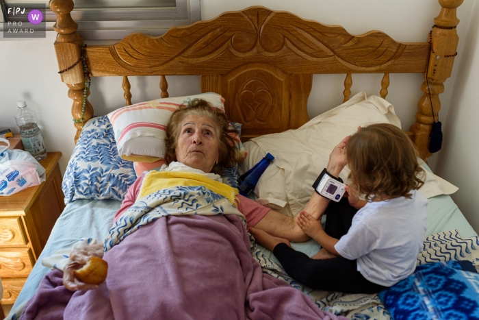 Photo by: <a href="https://www.fpja.com/documentary-family-photography-contests/emanuelle-rigoni/recife/2092401">Emanuelle Rigoni - Brazil</a> A little girl looks at her grandmother's wristband while she lays in bed in this documentary-style family photo captured by a Recife photographer.
