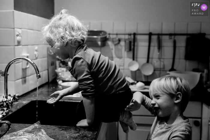 Photo by: <a href="https://www.fpja.com/documentary-family-photography-contests/angeline-dobber-jansen/gelderland/2093501">Angeline Dobber-Jansen - Netherlands</a> A little girl sits on a countertop playing with the sink faucet while her brother makes sure she doesn't fall in this picture captured by a Gelderland, Netherlands family photojournalist.