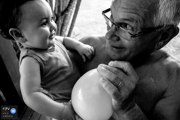 Photo by: <a href="https://www.fpja.com/documentary-family-photography-contests/thiago-braga/florianopolis/2093711">Thiago Braga - Brazil</a> A grandfather blows up a balloon for his granddaughter in this family picture by a Florianopolis photographer.