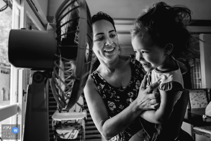 Une mère tient sa fille devant un fan sur cette photo enregistrée par un photographe de famille de style documentaire primé au Brésil, Rio Grande do Sul.