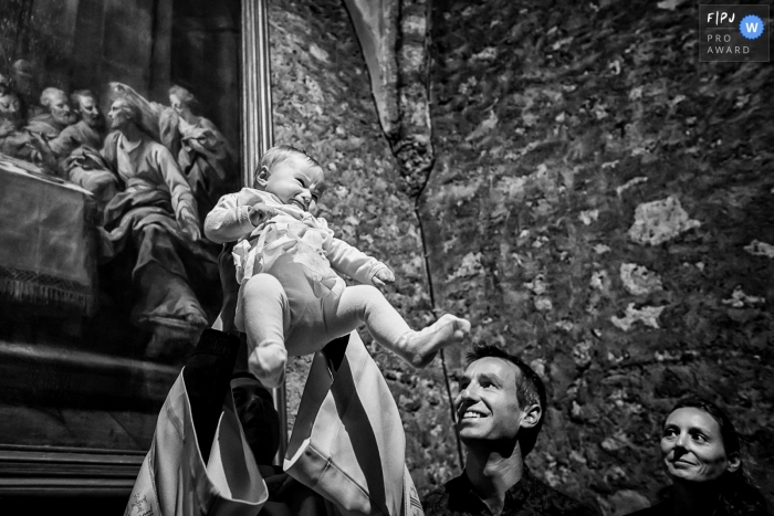 Un prêtre soulève un bébé dans les airs sous le regard des parents sur cette photo réalisée par un photojournaliste de famille français.