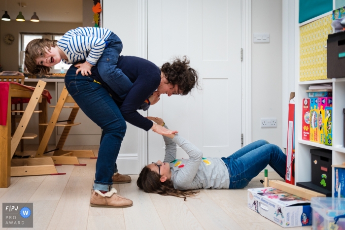 Photo by: <a href="https://www.fpja.com/documentary-family-photography-contests/alice-chapman/cambridgeshire/2096026">Alice Chapman - United Kingdom</a> A mother bends down to her daughter who lays on the ground while her son holds onto her back in this documentary-style family photo captured by a Cambridgeshire, England photographer.