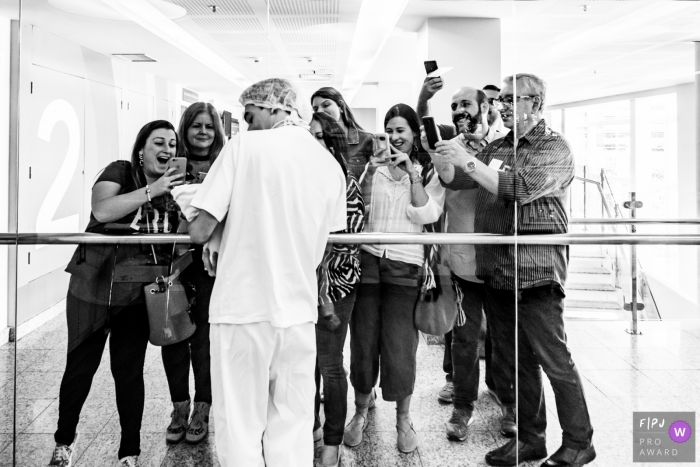 Photo by: <a href="https://www.fpja.com/documentary-family-photography-contests/liliane-cotta/rio-de-janeiro/2096141">Liliane Cotta - Brazil</a> Family members take pictures through a large hospital window as a nurse holds up a newborn for them to see in this black and white birth photo composed by a Rio de Janeiro, Brazil photographer.
