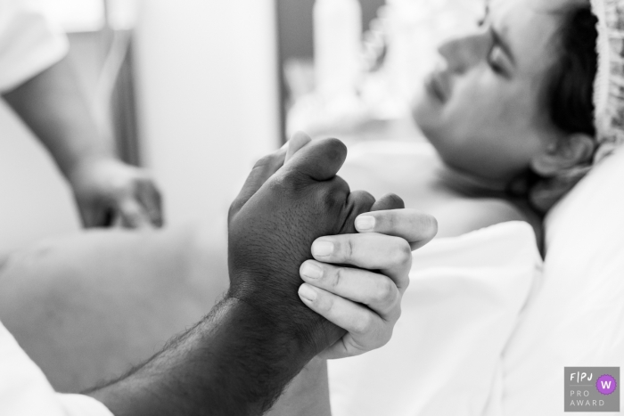 Photo by: <a href="https://www.fpja.com/documentary-family-photography-contests/liliane-cotta/rio-de-janeiro/2096201">Liliane Cotta - Brazil</a> A husband holds his wife's hand in the hospital as she gives birth in this black and white image recorded by a documentary-style Rio de Janeiro, Brazil birth photographer.