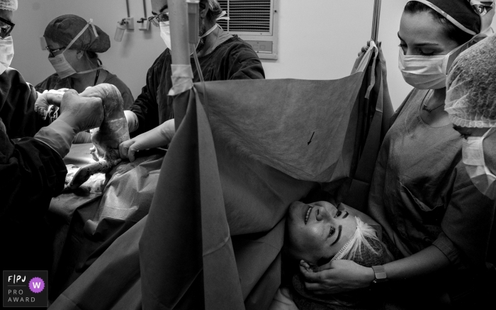 Photo by: <a href="https://www.fpja.com/documentary-family-photography-contests/lisandro-castro/cuiab%C3%A1/2096336">Lisandro Castro - Brazil</a> Nurses stand by a mother as the doctor pulls her baby out during a C-section in this black and white photo composed by a Cuiaba birth photographer.