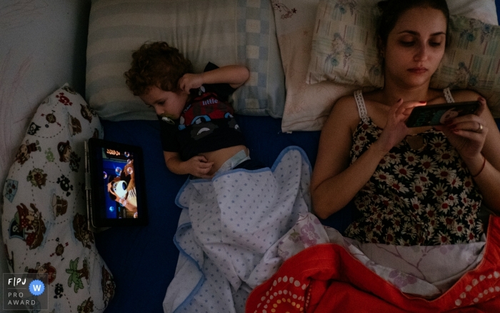 Un petit garçon regarde une émission sur un iPad pendant que sa mère regarde son téléphone et que les deux sont couchées au lit dans cette image créée par un photographe de la famille Cuiaba.