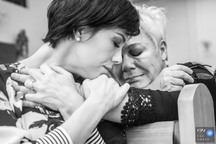 Photo by: <a href="https://www.fpja.com/documentary-family-photography-contests/ta%C3%ADs-motta/campinas/2096721">Taís Motta - Brazil</a> Two women hug each other over the back of a pew in this documentary-style family image recorded by a Campinas photographer.