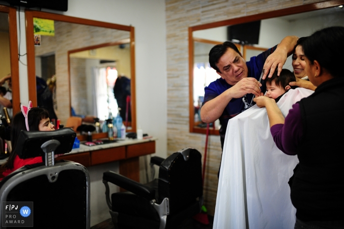 Photo by: <a href="https://www.fpja.com/documentary-family-photography-contests/nana-bonorino/rio-grande-do-sul/2097041">Nana Bonorino - Brazil</a> A family stands around a little boy as he gets his hair cut for the first time in this Family Photojournalist Association awarded photo by a Rio Grande do Sul, Brazil documentary family photographer.
