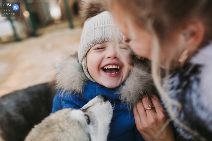 Photo by: <a href="https://www.fpja.com/documentary-family-photography-contests/elena-petrova/saint-petersburg/2097431">Elena Petrova - Russia</a> A mother holds her son while he laughs as a dog sniffs him in this documentary-style family image recorded by a Saint Petersburg, Russia photographer.