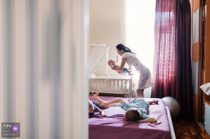 Photo by: <a href="https://www.fpja.com/documentary-family-photography-contests/elena-petrova/saint-petersburg/2097456">Elena Petrova - Russia</a> A mother lifts her baby out of his crib while her two other sons play on a bed in this Family Photojournalist Association contest awarded photo created by a Saint Petersburg, Russia family photographer.