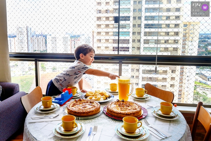 A little boy reaches across a table for the pitcher of orange juice in this award-winning photo by a Goiania family photographer.