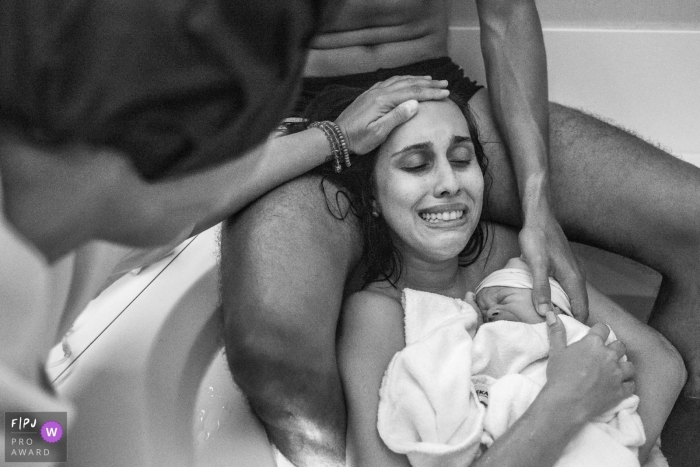 A husband supports his wife as she holds their newborn baby after her at-home water birth in this black and white photo by a Rio de Janeiro, Brazil birth photographer. 