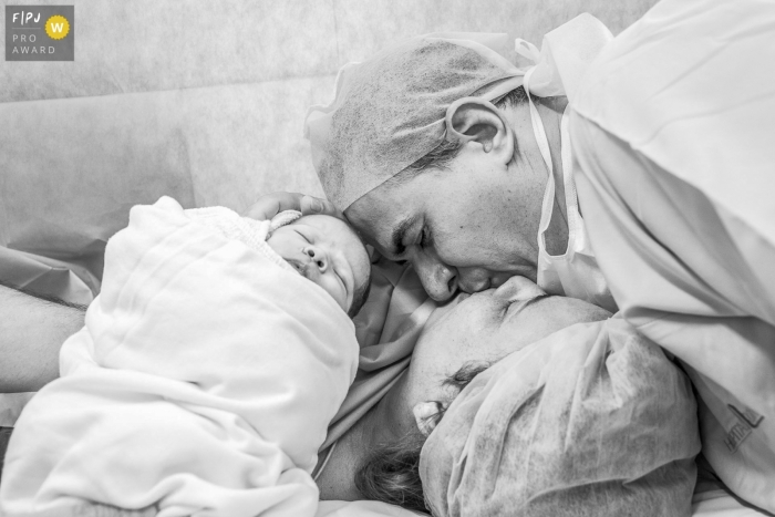 Photo by: <a href="https://www.fpja.com/documentary-family-photography-contests/claudia-ruiz/rio-de-janeiro/2097896">Claudia Ruiz - Brazil</a> A husband kisses his wife as they hold their baby in the hospital in this birth photo captured by an award-winning Rio de Janeiro, Brazil photographer.