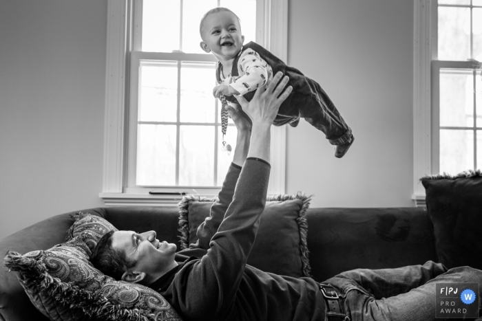 A father lays on the couch and holds his baby boy up in the air in this family picture by a Connecticut photographer. 