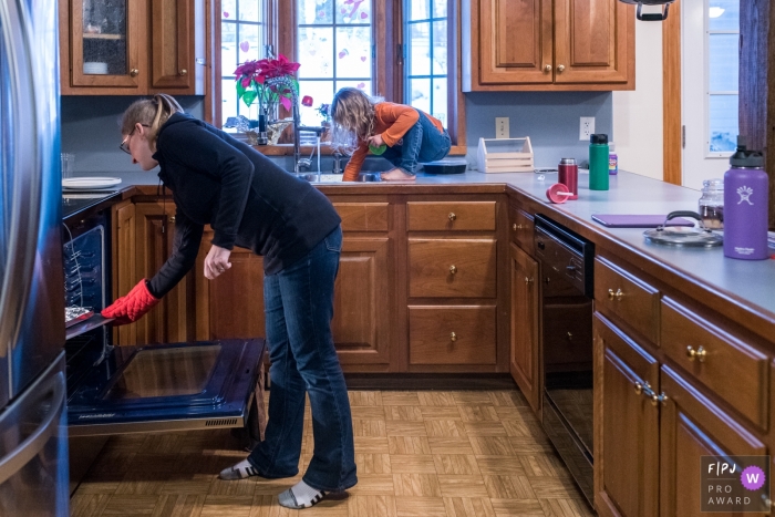 Une petite fille est assise sur le comptoir de la cuisine en train de jouer dans l'évier pendant que sa mère met de la nourriture au four sur cette photo prise par un photographe de la famille du Connecticut.