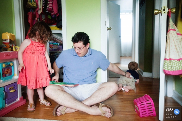 Photo by: <a href="https://www.fpja.com/documentary-family-photography-contests/katie-mcmenamin/philadelphia/2098716">Katie McMenamin - United States</a> A father holds onto his son who tries to crawl out of the room as he tries to read to his daughter in this Family Photojournalist Association contest awarded photo created by a Philadelphia family photographer.