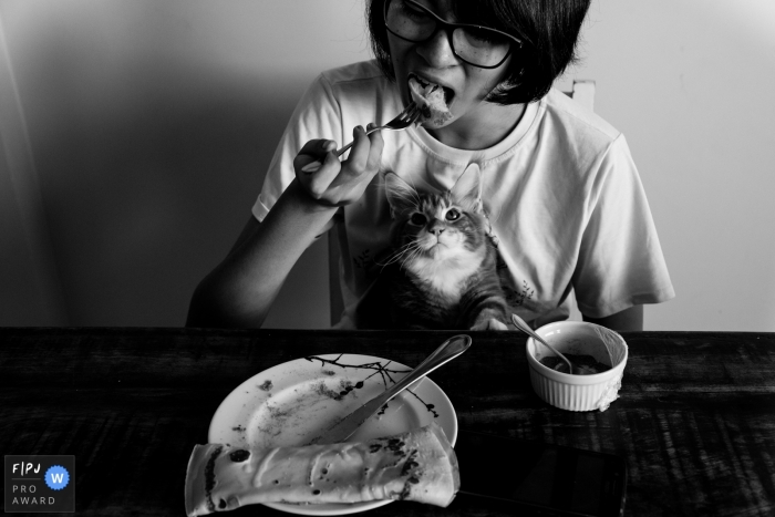 Photo by: <a href="https://www.fpja.com/documentary-family-photography-contests/carolina-belini/goiania/2099251">Carolina Belini - Brazil</a> Goiana, Brazil Family Photography | Child sitting at a table eating with a cat in their lap.
