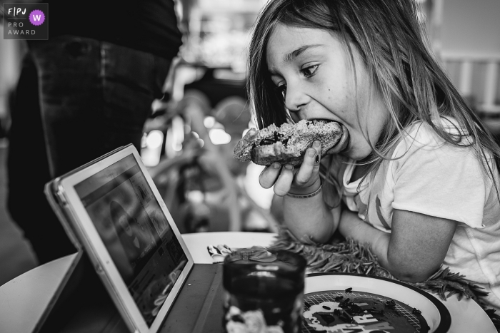 Photo by: <a href="https://www.fpja.com/documentary-family-photography-contests/hiske-boon/noord-brabant/2099542">Hiske Boon - Netherlands</a> Noord Brabant family photography | Black and white photo of a young girl watching an iPad as she eats a meal