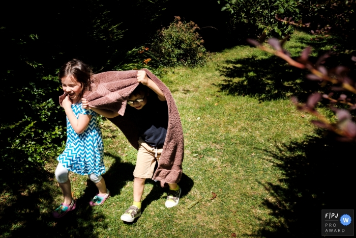 Photo by: <a href="https://www.fpja.com/documentary-family-photography-contests/jennifer-adams/gloucestershire/2099634">Jennifer Adams - United Kingdom</a> Gloucestershire Family Photograph of siblings running through the garden with a towel over their heads.