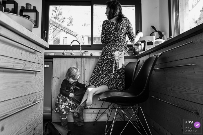 Photo by: <a href="https://www.fpja.com/documentary-family-photography-contests/nadine-court/savoie/2099684">Nadine Court - France</a> Savoie Family Photography of a young girl taking her mom's boot off during the kitchen chores.