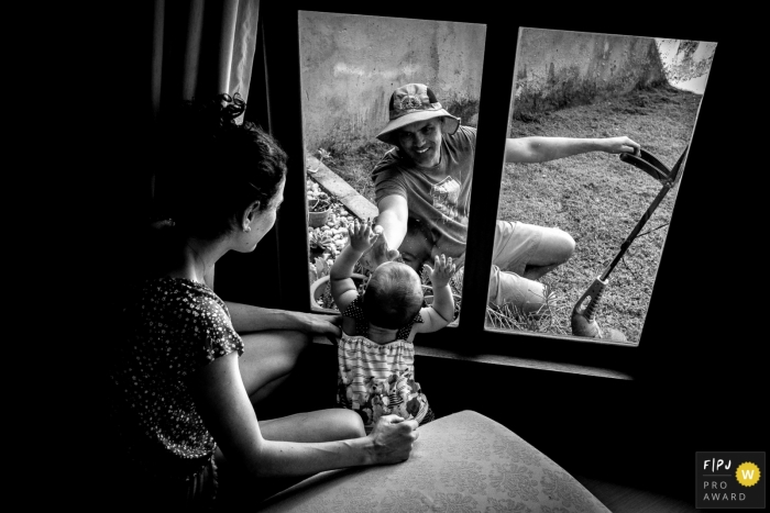 Florianpolis Family Photo | Father mowing lawn in the home garden connects with his daughter who is on his mother's lap at a window in the home room.