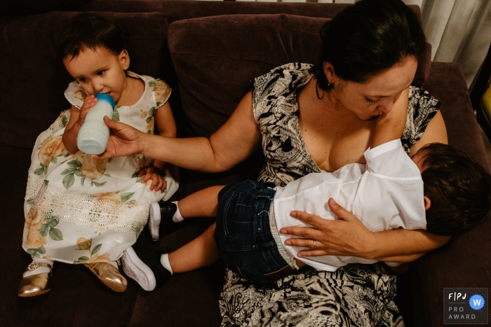 Photo by: <a href="https://www.fpja.com/documentary-family-photography-contests/thiago-braga/florianopolis/2099899">Thiago Braga - Brazil</a> Santa Catarine Family Photography | mother with her two sons, twins, breastfeeding one and bottle feeding the other, after the end of the 2 year birthday party on the couch.
