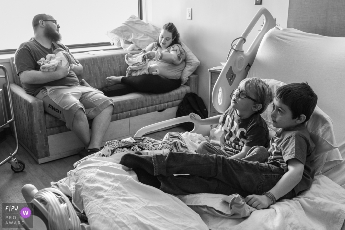 Photographie de famille et de naissance du Connecticut | La nouveauté de leurs nouvelles sœurs jumelles s'est dissipée et deux grands frères occupent le lit d'hôpital de maman pour regarder la télévision pendant que maman et papa s'occupent des bébés en arrière-plan.