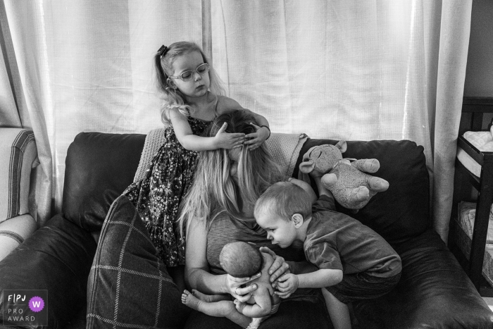 Photo by: <a href="https://www.fpja.com/documentary-family-photography-contests/ang-waterton/ontario/2100252">Ang Waterton - Canada</a> Ontario mother holds baby while boy reaches to kiss baby and girl styles mother's hair | Canada family photojournalism session.
