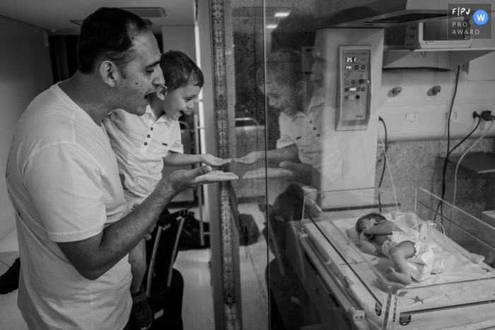 Documentary birthing photography in Recife - Pernambuco dad and big brother see newborn baby behind glass.