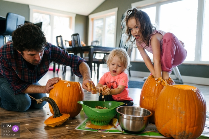 Photo by: <a href="https://www.fpja.com/documentary-family-photography-contests/viara-mileva/kingston/2101282">Viara Mileva - Canada</a> Family carves pumpkins as girl climbs into pumpkin | Kingston Family Photography