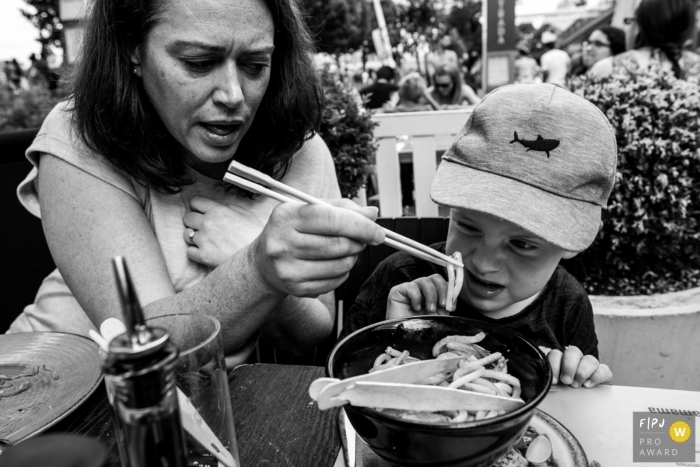 Photo by: <a href="https://www.fpja.com/documentary-family-photography-contests/raluca-chase/essex/2101451">Raluca Chase - United Kingdom</a> England family photoshoot: Mom trying to feed her son in at a Wagamama restaurant in London