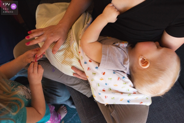 Photo by: <a href="https://www.fpja.com/documentary-family-photography-contests/amber-roy/state-college/2101694">Amber Roy - United States</a> Mother having her nails done by young daughter as she nurses her infant | State College Family Photography