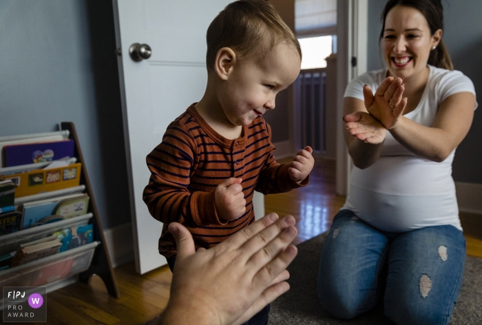 Photo by: <a href="https://www.fpja.com/documentary-family-photography-contests/ang-waterton/ontario/2101836">Ang Waterton - Canada</a> Ontario toddler makes face while dancing as parents clap - Canada family photo session at home.