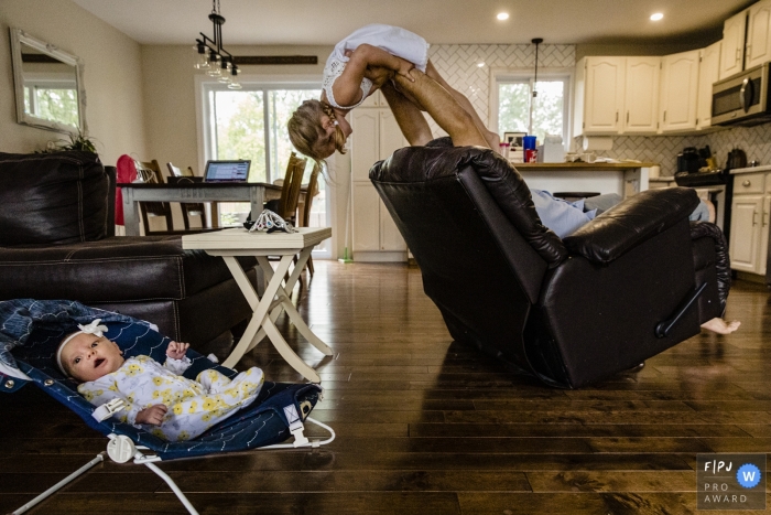 Photo by: <a href="https://www.fpja.com/documentary-family-photography-contests/ang-waterton/ontario/2101850">Ang Waterton - Canada</a> Ontario, Canada family photography - baby looks surprised while dad holds daughter in air in background