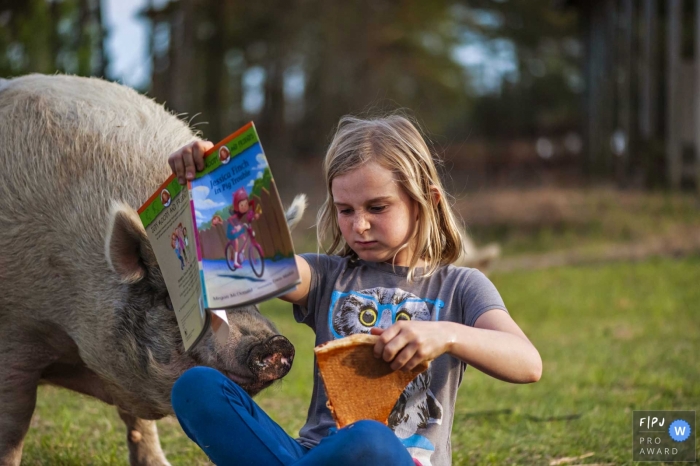 Photo by: <a href="https://www.fpja.com/documentary-family-photography-contests/brooke-mayo/outer-banks/2101941">Brooke Mayo - United States</a> Girl reading a book and eating pizza has an inquisitive pig visitor looking for a snack | North Caroliina Family Photography