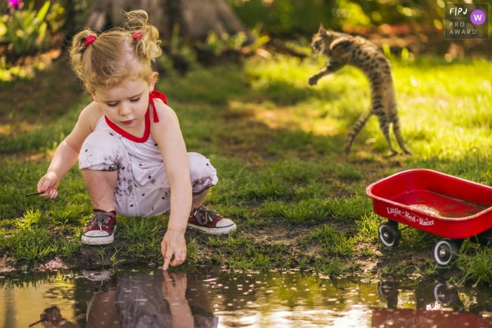 Young girl plays in a puddle with her little red wagon and a cat leaping behind her | Outer Banks Family Photography