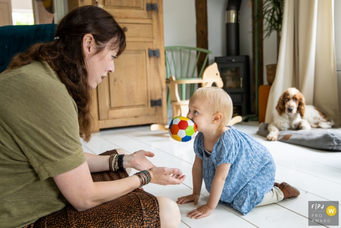 Photo by: <a href="https://www.fpja.com/documentary-family-photography-contests/angeline-dobber/gelderland/2102040">Angeline Dobber - Netherlands</a> Crawling toddler brings mom a ball in her mouth as dog watches | Netherlands Family Session