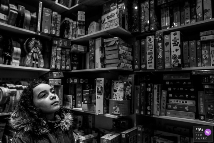 Photo by: <a href="https://www.fpja.com/documentary-family-photography-contests/nadine-court/savoie/2102156">Nadine Court - France</a> Auvergne-Rhone-Alpes family photo session in town | A girl dreams in a toy store with lots of board games