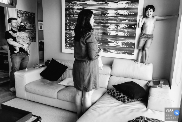 Photo by: <a href="https://www.fpja.com/documentary-family-photography-contests/alexandre-tessler/porto-alegre/2102225">Alexandre Tessler - Brazil</a> Boy stands on the back of sofa as mom coaxes him to get dressed | Porto Alegre Family Photographer
