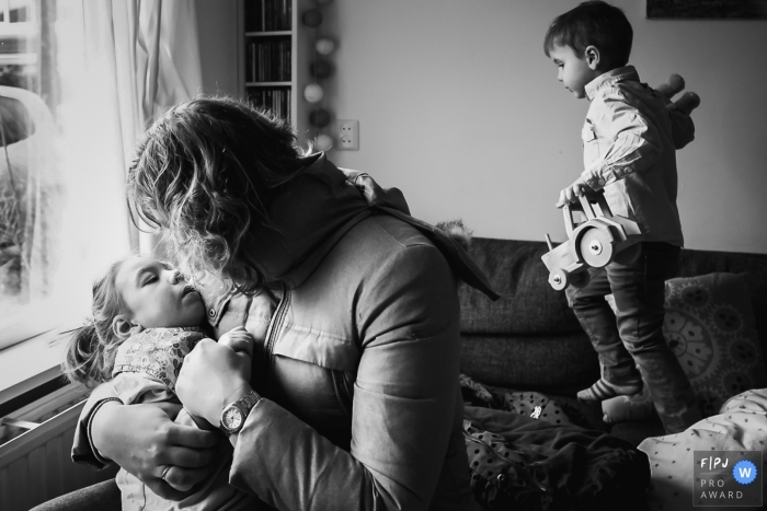 Photo by: <a href="https://www.fpja.com/documentary-family-photography-contests/marjolijn-maljaars/zuid-holland/2102238">Marjolijn Maljaars - Netherlands</a> A mother is hugging her child as a young boy walks across the sofa with a toy tractor | Zuid Holland Family Photography
