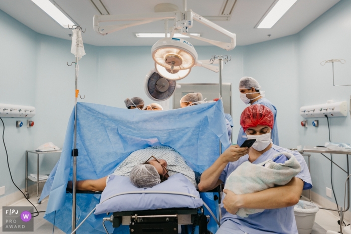 Rio de Janeiro Birth Photographer | Photo of girl being born by c-section, with the father taking the first selfie with her