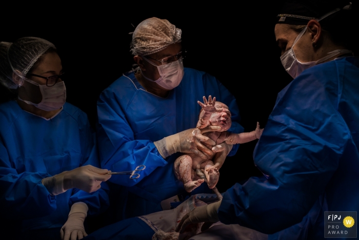 Newborn reaches outwards with arms as it is pulled up by a doctor | Brazil Birth Photography Session