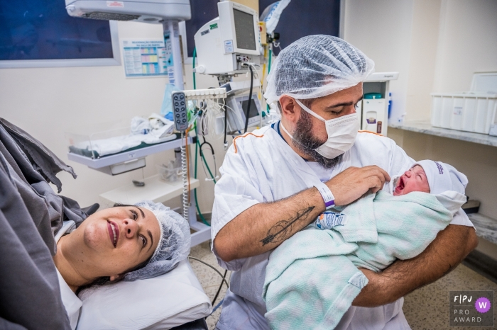 Photo by: <a href="https://www.fpja.com/documentary-family-photography-contests/ta%C3%ADs-motta/campinas/2102404">Taís Motta - Brazil</a> Father holds yawning newborn swaddled in blankets as mom looks on | Sao Paulo Birthing