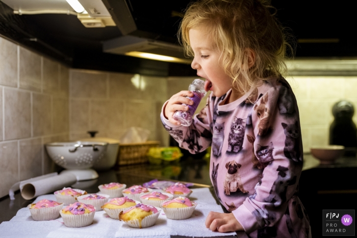 Young girl in the kitchen a jar of sprinkles to her mouth with a spread of cupcakes on the counter before her | Noord Holland Family Session