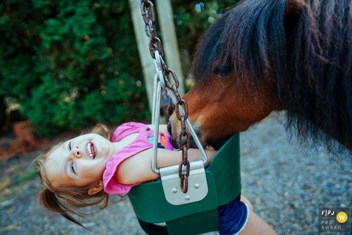 Photo by: <a href="https://www.fpja.com/documentary-family-photography-contests/ron-storer/seattle/2102752">Ron Storer - United States</a> Washington family photo of a mini horse tickling child on swing